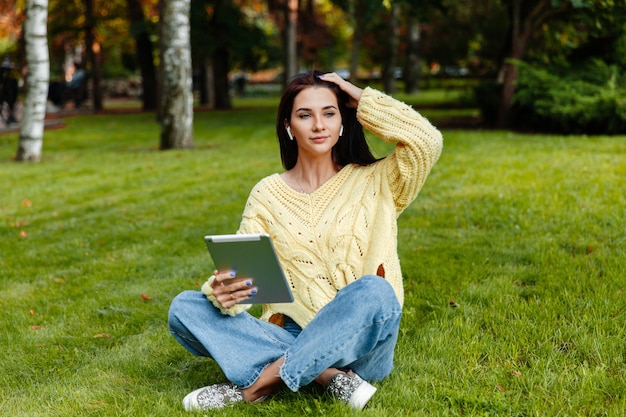 Das Mädchen sitzt im Park und beschäftigt sich mit dem iPad. junge brünette sitzt auf dem gras und schaut in die tablette. Mädchen im Herbstpark.