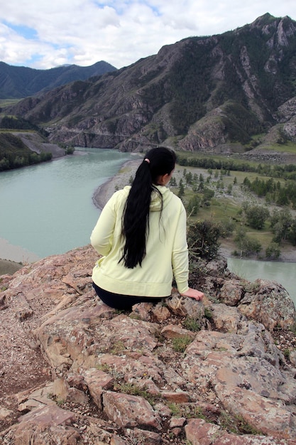 Das Mädchen sitzt mit dem Rücken zu uns am Rand des Berges vor dem Hintergrund eines blauen Flusses, Berge und Wald