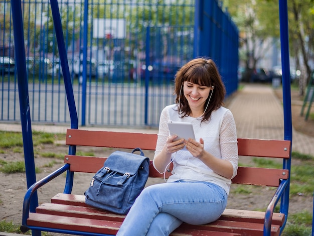 Das Mädchen sitzt mit einer Tablette auf einer Bank und lacht. Fernkommunikation in Quarantäne. Brünettes Mädchen in einer weißen Jacke und Jeans auf der Straße