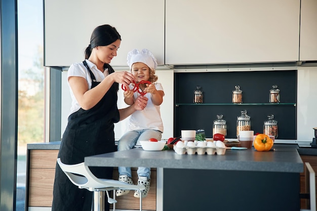 Foto das mädchen trägt den haupthut. mutter und ihre tochter bereiten essen in der küche vor.