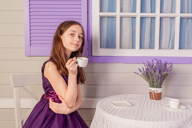 Das Mädchen trinkt Tee aus einer weißen Tasse. Ein junges Mädchen in einem lila Kleid am Tisch trinkt Tee.