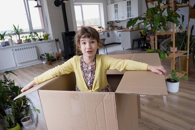 Das Mädchen versteckt sich in einer großen Kiste und sieht komisch aus in einem Raum innerhalb des Hauses. Umzug in ein neues Haus, Auspacken von Dingen, Mieten einer Wohnung, Hypothekenwohnungsproblem