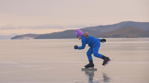 Das Mädchen Zug auf Eisschnelllauf Das Kind Schlittschuhe im Winter in blauer Sportbekleidung Sportbrille Kinder-Eisschnelllauf-Sport Zeitlupe im Freien Schneebedeckte Berge schönes Eis