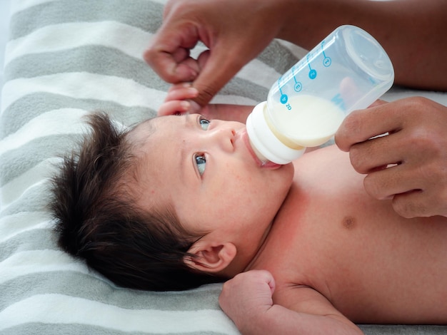Das Porträt der asiatischen Mutter füttert das australische asiatische dreiwöchige Neugeborene mit Milchformel aus einem Flaschenkonzept von Mutterschaft und Kindheit oder Kleinkind, das auf dem weißen Bett liegt