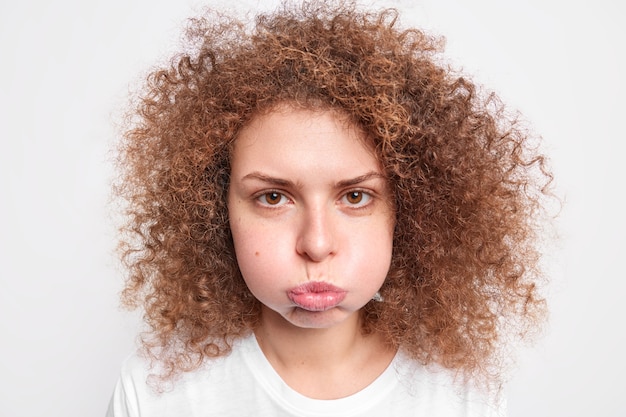 Foto das porträt der beleidigten, lockigen, schönen jungen frau bläst wangen hat einen unzufriedenen ausdruck, der negative emotionen ausdrückt, die über der weißen wand isoliert sind menschliche gesichtsausdrücke und gefühle