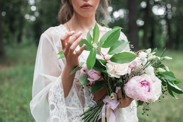 Das Porträt der Braut im Freien in einem weißen Spitzen-Peignoir hält in ihren Händen einen Hochzeitsstrauß mit Rosen