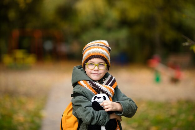 Das Porträt des Jungen in den Gläsern und in der warmen Strickmütze und im Schal mit seinem Spielzeug im Herbstpark