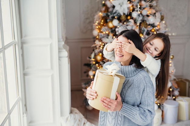 Das Porträt des kleinen Mädchens schließt die Augen der Mutter und gratuliert ihr zum Neujahrs- oder Weihnachtsstand in der Nähe des Fensters im Wohnzimmer