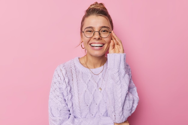 Das Porträt einer angenehm aussehenden Frau lächelt breit und zeigt perfekte weiße Zähne, die die Hand am Rand einer Brille hält, die in einem Strickpullover gekleidet ist, der über rosa Hintergrund isoliert ist. Menschen und positive Emotionen