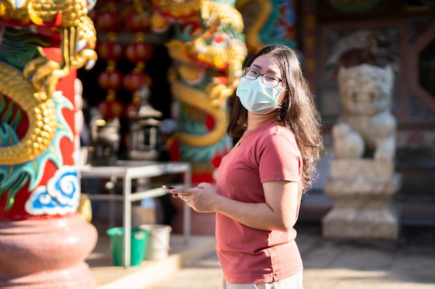 Das Porträt einer asiatischen Reisenden, die Schutzmaskenkeime trägt, sucht Informationen auf dem Smartphone im chinesischen buddhistischen Tempel oder Schrein, Prävention der Ausbreitung des COVID-19-Virus, Chinesisches Neujahr