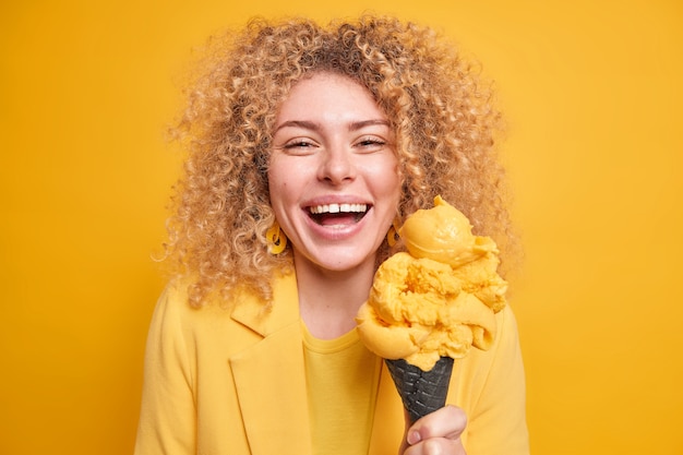 Das Porträt einer fröhlichen Frau mit lockigem, buschigem Haar lächelt im Großen und Ganzen hat Spaß und genießt es, appetitliches Kegeleis zu essen, das in gelber Kleidung gekleidet ist, und drückt positive Emotionen aus. Sommer-Lifestyle-Konzept