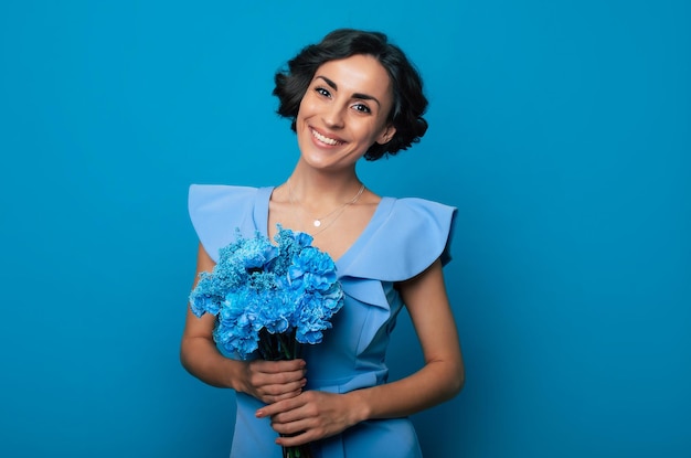 Das Porträt einer glücklichen, aufgeregten, wunderschönen jungen Frau in einem eleganten blauen Kleid posiert mit einem frischen Strauß blauer Blumen. Muttertag. Feiertage für Frauen. Frühling. Frauenrechte