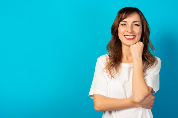 Das Porträt einer jungen freundlichen Frau im lässigen T-Shirt hält ihre Hände unter ihrem Kinn und lächelt auf Blau