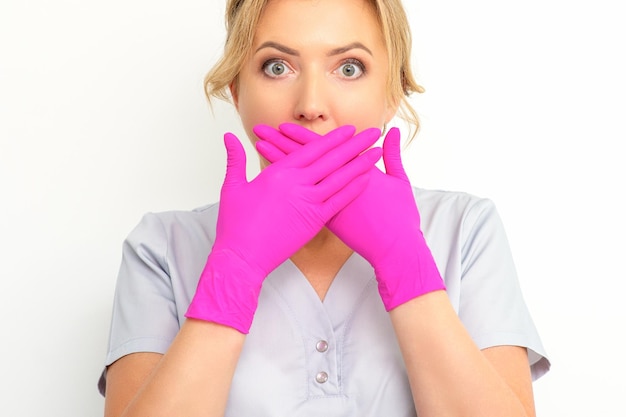 Das Porträt einer jungen kaukasischen Ärztin oder Krankenschwester ist schockiert und bedeckt ihren Mund mit ihren rosa behandschuhten Händen vor einem weißen Hintergrund