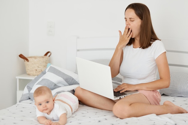 Das Porträt einer müden, schläfrigen Frau, die ein weißes T-Shirt trägt und mit ihrer kleinen Tochter auf dem Bett sitzt und online arbeitet, einen Laptop hält und gähnt, online zu arbeiten, fühlt sich schläfrig an