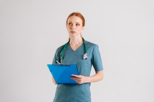 Das Porträt einer nachdenklichen jungen Ärztin in medizinischer Uniform, die ein Rezept in die Zwischenablage schreibt, füllt die Krankengeschichte aus, die auf weißem, isoliertem Hintergrund im Studio wegschaut