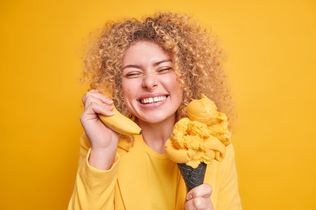 Das Porträt einer positiven kaukasischen Frau lächelt breit und zeigt weiße, gleichmäßige Zähne, die in guter Stimmung sind, während sie das Lieblingsdessert isst, eine Banane in der Nähe des Ohrs hält, die Augen vor dem Vergnügen schließt, isoliert über der gelben Wand