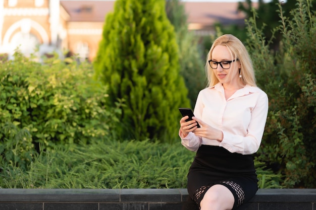 Das Porträt einer schönen, fröhlichen Geschäftsfrau in Hemd und schwarzem Rock mit ihrem Smartphone sitzt vor dem Hintergrund eines Blumenbeets