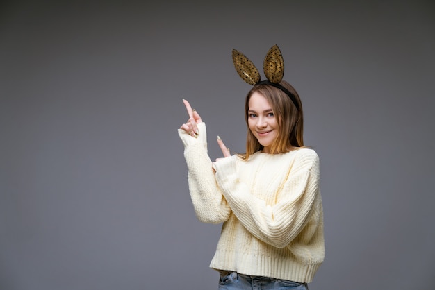 Das Porträt einer schönen fröhlichen jungen Frau in einem Pullover und in Hasenohren zeigt ihren Finger zur Seite an einer grauen Wand