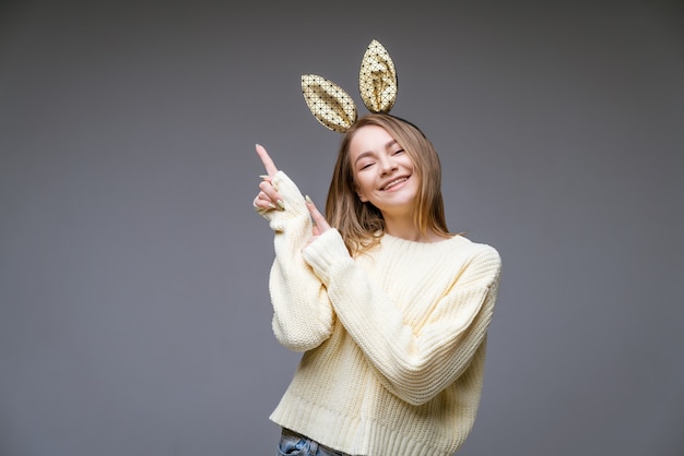 Das Porträt einer schönen fröhlichen jungen Frau in einem Pullover und in Hasenohren zeigt ihren Finger zur Seite auf Grau