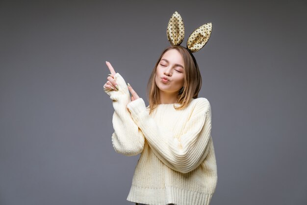 Das Porträt einer schönen fröhlichen jungen Frau in einem Pullover und in Hasenohren zeigt ihren Finger zur Seite