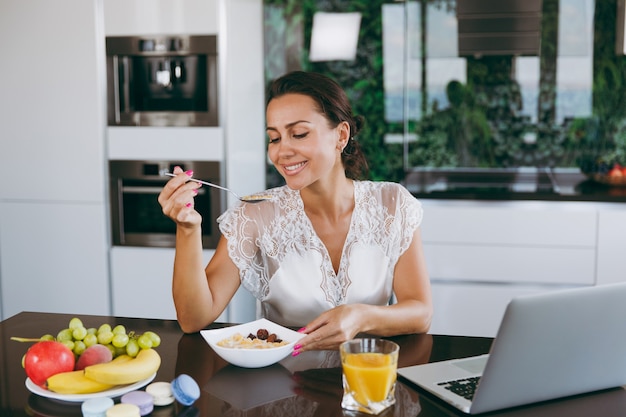 Das Porträt einer schönen glücklichen Frau, die beim Frühstück mit Müsli und Milch mit Laptop arbeitet