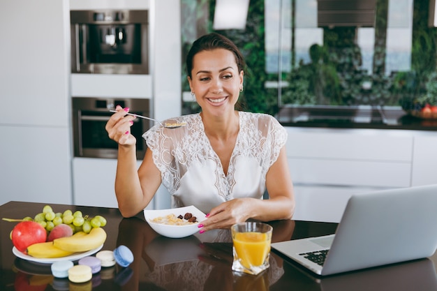 Das Porträt einer schönen glücklichen Frau, die beim Frühstück mit Müsli und Milch mit Laptop arbeitet