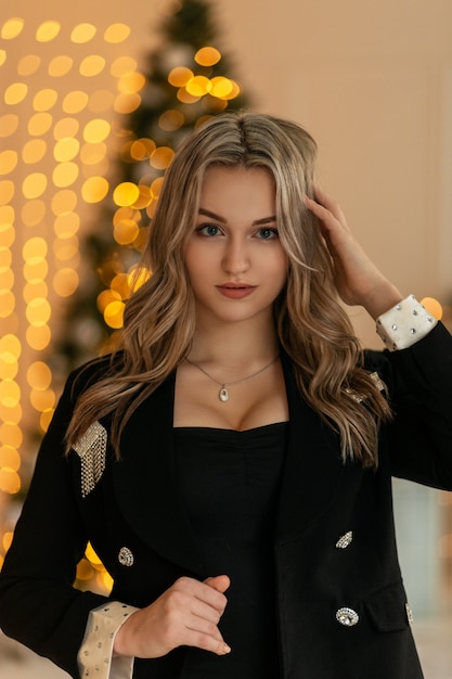 Das Porträt einer schönen jungen Frau in eleganter schwarzer Kleidung mit einem Blazer mit schönen üppigen Brüsten sieht die Kamera auf einem Hintergrund von Weihnachtsdekorationen und gelben Bokeh-Lichtern an