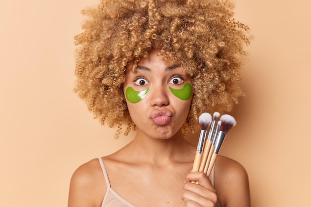 Das Porträt einer überraschten Frau mit lockigem, buschigem Haar hält die Lippen gefaltet und blickt in die Kamera. Sie hält Kosmetikpinsel, die Make-up auftragen werden, und trägt ein lässiges T-Shirt, das über beigem Hintergrund isoliert ist