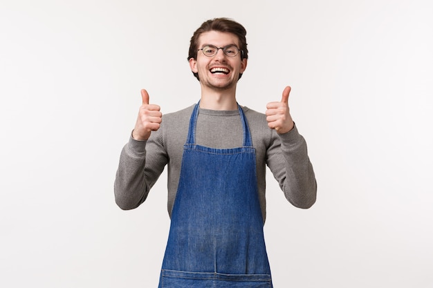 Das Porträt eines aufgeregten und optimistischen positiven jungen Mannes in der Schürze, der als Barista im Café oder Barkeeper arbeitet, zeigt Daumen hoch in der Zustimmung, wie etwas Fantastisches, garantiert guten Geschmack