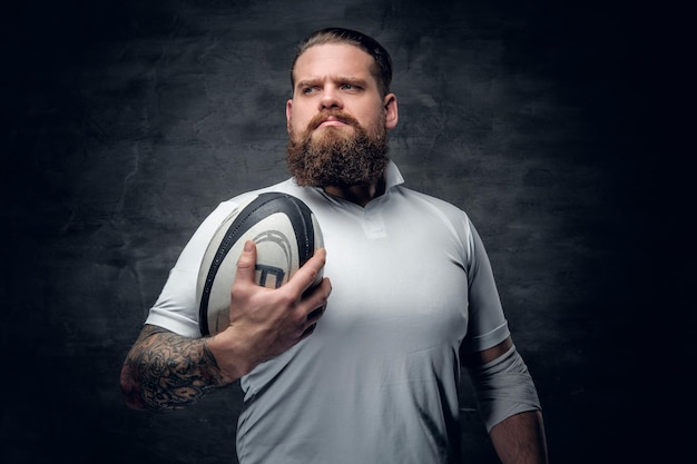 Das Porträt eines bärtigen Rugbyspielers mit Tattoos auf seinen Armen hält einen Spielball.