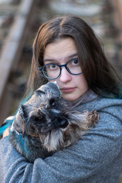 Das Porträt eines brünetten Mädchens in einem grauen Mantel hält einen Zwergschnauzer in den Armen. Der Zwergschnauzer und das Mädchen schauen dich an. Lieblingshaustierkonzept