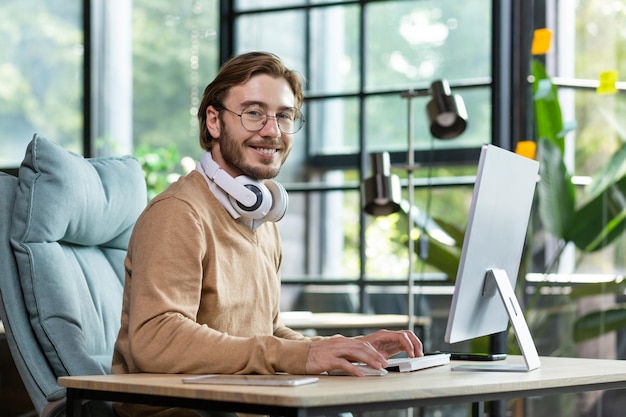 Das Porträt eines jungen freiberuflichen Programmierers sitzt mit weißen Kopfhörern im Büro und arbeitet an der