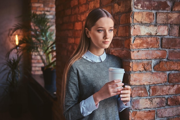 Das Porträt eines jungen Mädchens in einem eleganten grauen Kleid, das sich an eine Ziegelwand lehnt, hält eine Tasse Kaffee zum Mitnehmen und schaut weg.