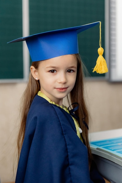 Das Porträt eines Mädchen der ersten Klasse in einem festlichen Hut, das für ein Foto an einem Schreibtisch in einem Klassenzimmer posiert
