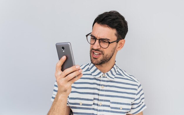 Das Porträt eines wütenden, bärtigen, gutaussehenden Geschäftsmannes trägt eine runde Brille und ein legeres Hemd blickt wütend in das Handy, das auf weißem Hintergrund der Studiowand posiert Menschen-Emotion-Technologie-Konzept