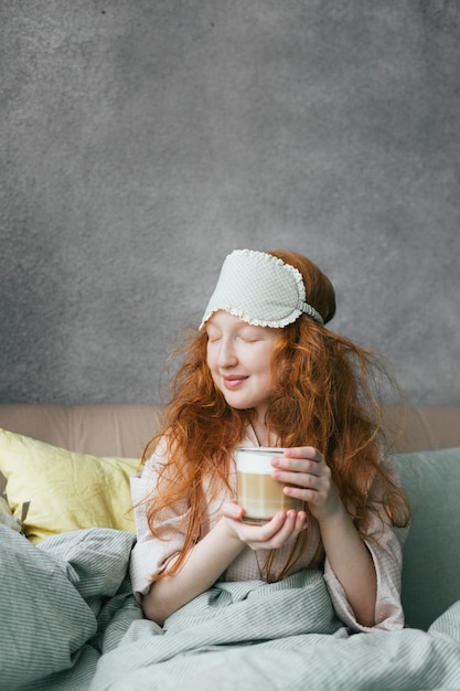 Das rothaarige süße Mädchen mit einer Tasse Kaffee genießt das Leben am frühen Morgen in ihrem Bett.