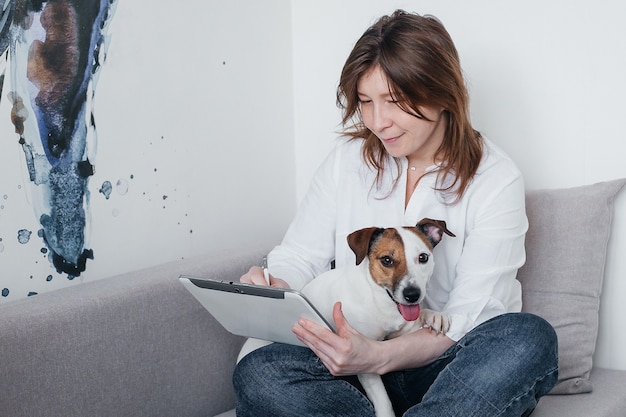 Das schöne Mädchen sitzt zu Hause mit einer Tablette in der Hand neben einem Jack Russell-Hund auf der Couch