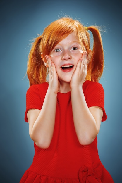 Das schöne Porträt eines überraschten kleinen Mädchens mit roten Haaren im roten Kleid auf Blau