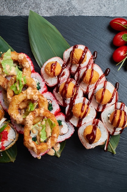 Das Set Sushi-Rollen liegt auf Bambusblättern mit Limette, Kräutern und roten Tomaten, die auf einem schwarzen Steinschiefer serviert werden.