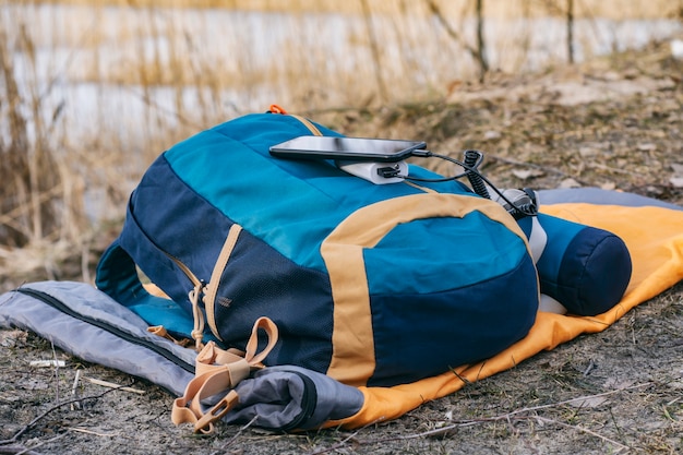 Foto das smartphone wird mit einem tragbaren ladegerät aufgeladen. power bank lädt das telefon im freien mit einem rucksack für den tourismus im hintergrund der natur und des flusses auf.