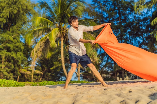 Das sommerliche Lifestyle-Porträt des Menschen bläst ein aufblasbares orangefarbenes Sofa am Strand der tropischen Insel auf. Entspannen und genießen Sie das Leben auf dem Luftbett