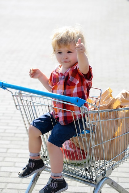 Das süße blonde Kind des kleinen Jungen im karierten Hemd sitzt im Einkaufswagen und isst an einem sonnigen Tag Kekse aus der Papierpackung im Freien