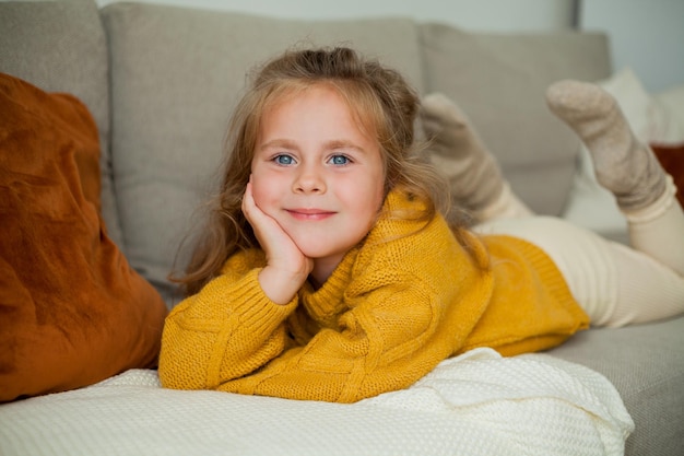 Das süße kleine blauäugige, lockige Mädchen, 4 Jahre alt, ruht auf dem Sofa in einem gemütlichen Haus Autumn Knitted