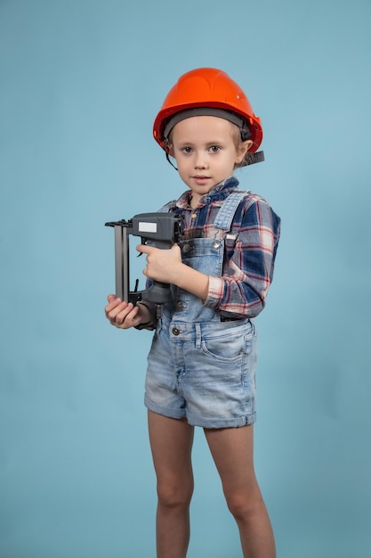 Das süße kleine kaukasische Kind trägt einen orangefarbenen Schutzhelm und hält einen Bauhefter an den Händen. Konzept für Bau und Reparatur.
