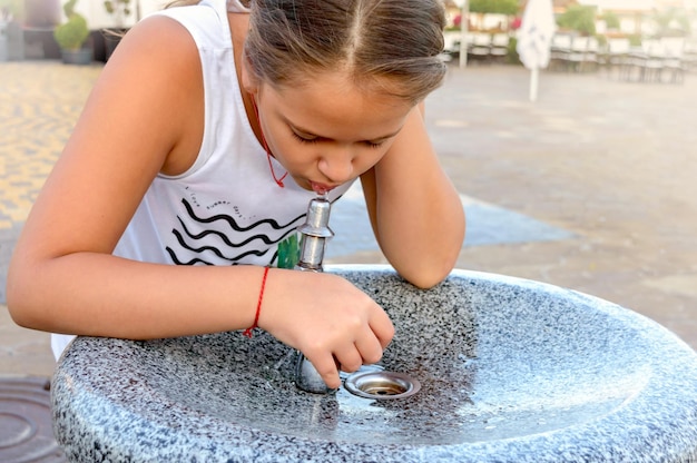 Das süße kleine Mädchen trinkt an einem heißen Sommertag Wasser aus dem öffentlichen Trinkwasserbrunnen