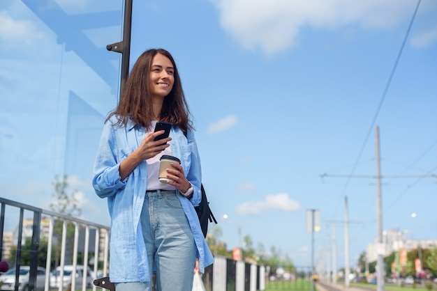 Das trendige Mädchen wartet morgens auf einen Bus oder eine Straßenbahn in der öffentlichen Verkehrsstation. Junge Frau mit Tasse Kaffee und Smartphone-Überwachungstransport durch die App.