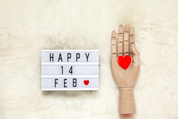 Das trendige menschliche Handmodell aus Holz hält ein rotes Herz und einen Leuchtkasten mit der Aufschrift Happy 14 FEB, was Valentinstag auf weißem Pelzteppich bedeutet.