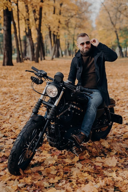 Das vertikale Bild eines durchdachten Bikers trägt eine Sonnenbrille, fährt Motorrad in Parkposen auf der Straße, trägt stilvolle Kleidung, genießt Freizeit und ein Hobby, bewundert die Natur, genießt sonniges Herbstwetter