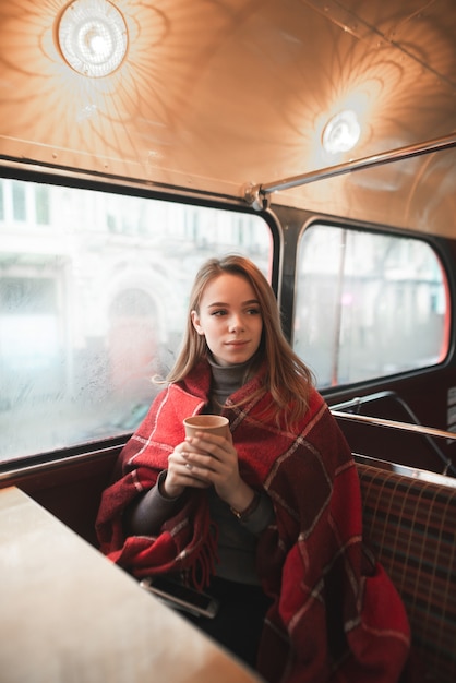 Das Winterporträt eines jungen Mädchens, bedeckt mit einer Decke, sitzt mit einer Tasse Kaffee am Fenster und schaut zur Seite